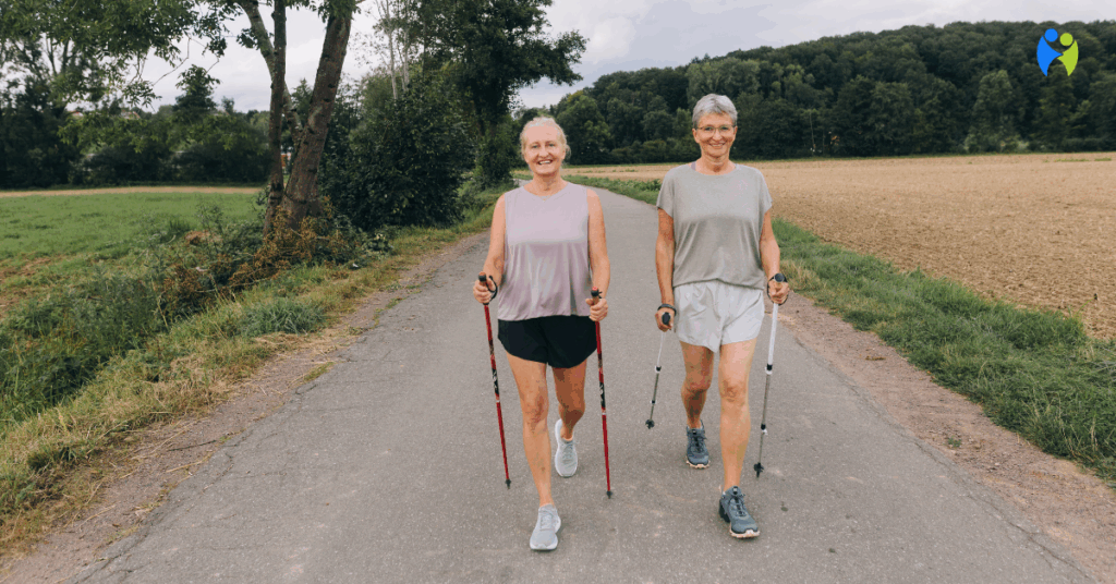 Dwie uśmiechnięte seniorki spacerujące z kijkami do nordic walking po wiejskiej drodze, otoczonej zielenią i polami.