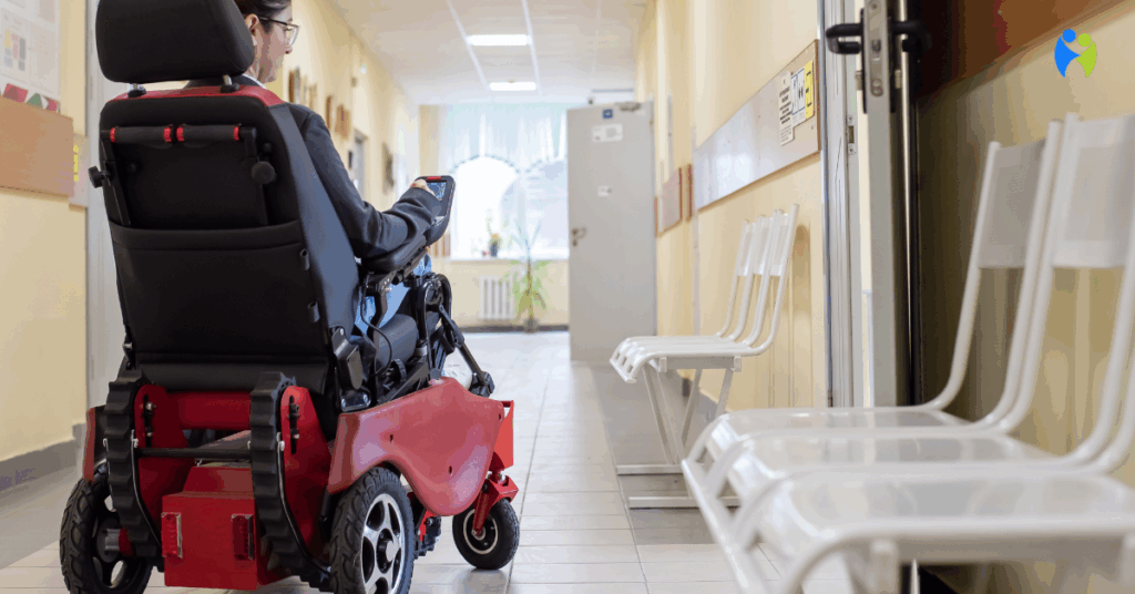 Osoba na czerwonym wózku inwalidzkim elektrycznym poruszająca się korytarzem placówki medycznej, widziana od tyłu.