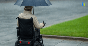 Osoba poruszająca się na wózku inwalidzkim elektrycznym jedzie w deszczu po miejskim chodniku, mając na sobie kurtkę przeciwdeszczową. Wózek osłonięty jest folią ochronną przed wilgocią.